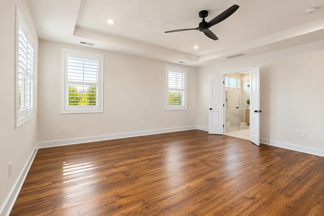 Empty bedroom with wooden-look flooring, a ceiling fan and an open door to an en-suite bathroom